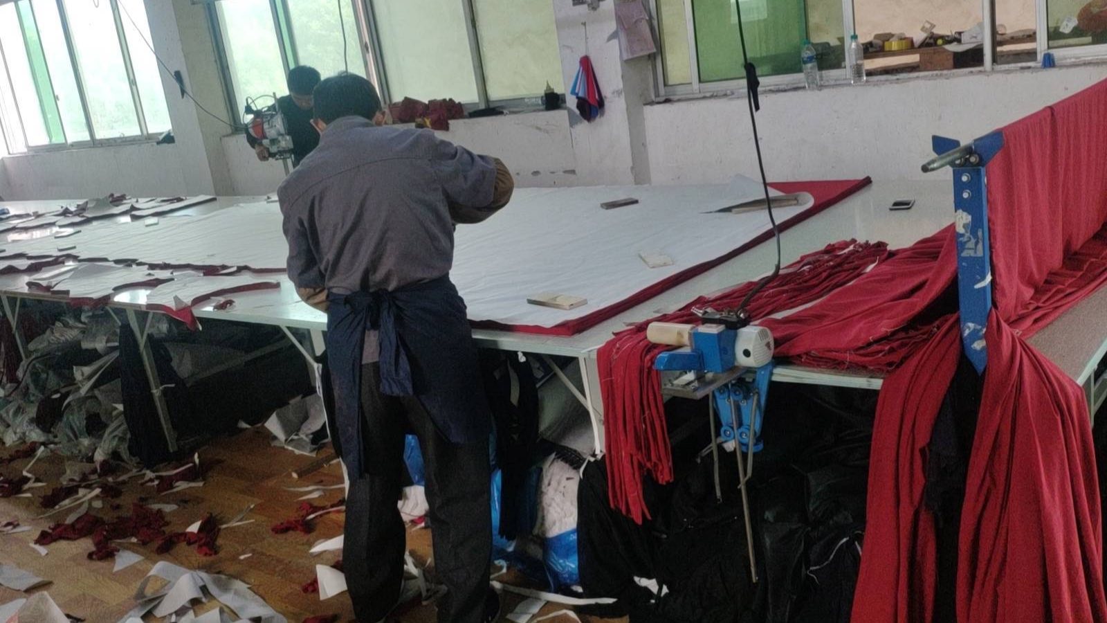 Workers spreading and cutting red fabric on a long cutting table with a round knife cutter
